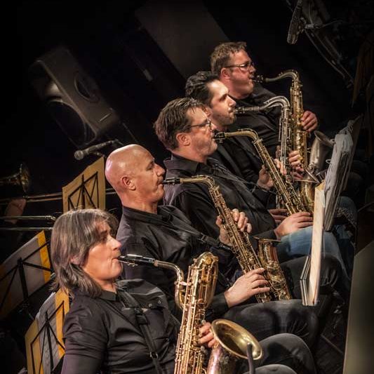 Vijf musici met saxofoons, gekleed in zwart, spelen samen op een podium.